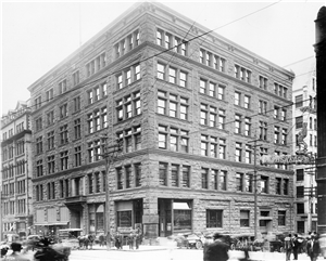 A six story brick building on a city corner. Pedestrians, horse and buggies, and early twentieth century automobiles pass by. 